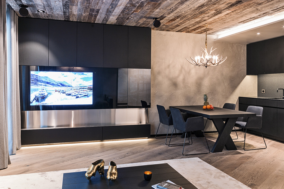 A vacation apartment in Switzerland with elegant furnishings, black dining table, fireplace, TV and antler-shaped chandelier.