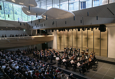 An orchestral performance in a modern concert hall in Andermatt, with an audience and a group of musicians on stage.