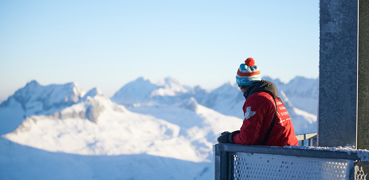 Eine Person in warmer Kleidung steht auf einem Balkon und schaut auf verschneite Berge in der Schweiz.