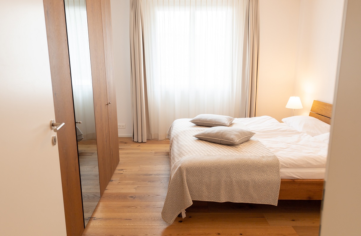 Andermatt Apartment bedroom with wooden bed frame, white bedding, beige throw blanket, gray pillows, bedside lamp, large curtained window, mirrored wardrobe, and warm light wood flooring.